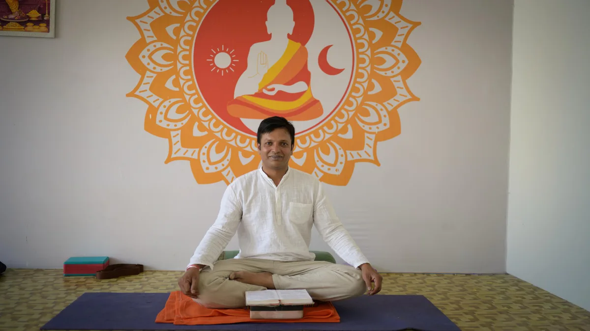 Somashekar, head teacher at Anantadrishti Yoga, seated cross-legged with an open book in front of a mandala mural in Rishikesh