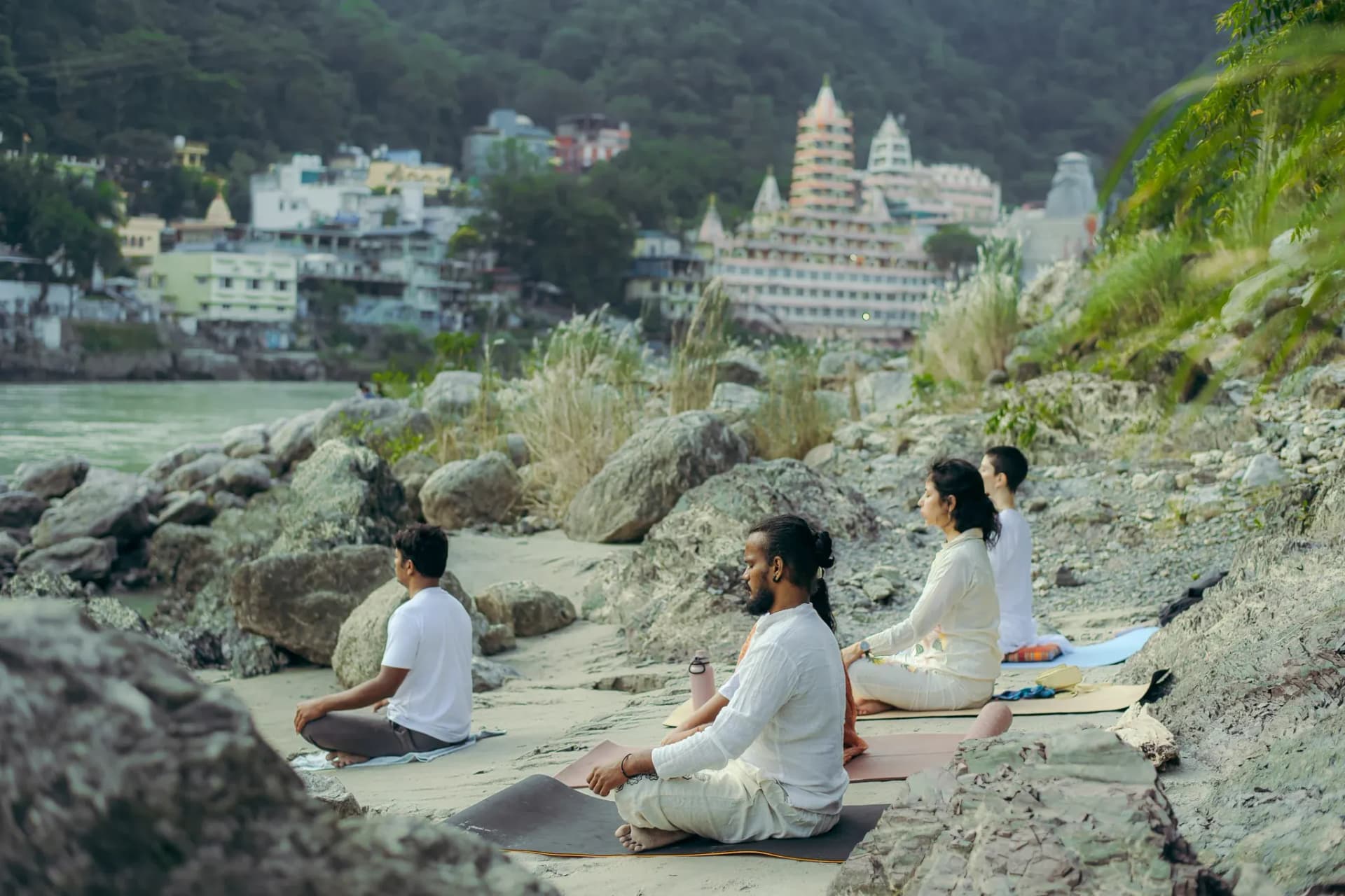 Students meditate cross-legged on the Ganga riverbank in Rishikesh with the Trimbakeshwar temple behind