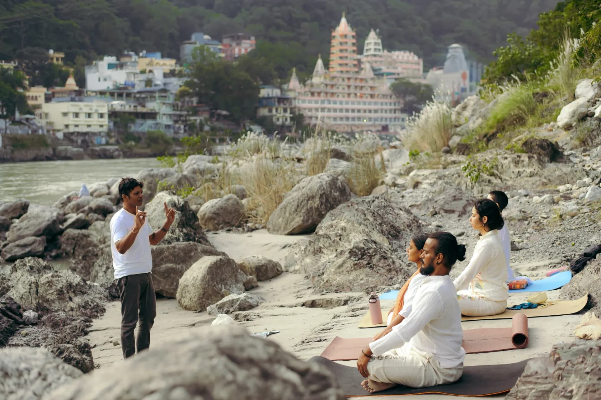 Yoga teacher instructs four seated students on the Ganga riverbank in Rishikesh, Trimbakeshwar temple behind