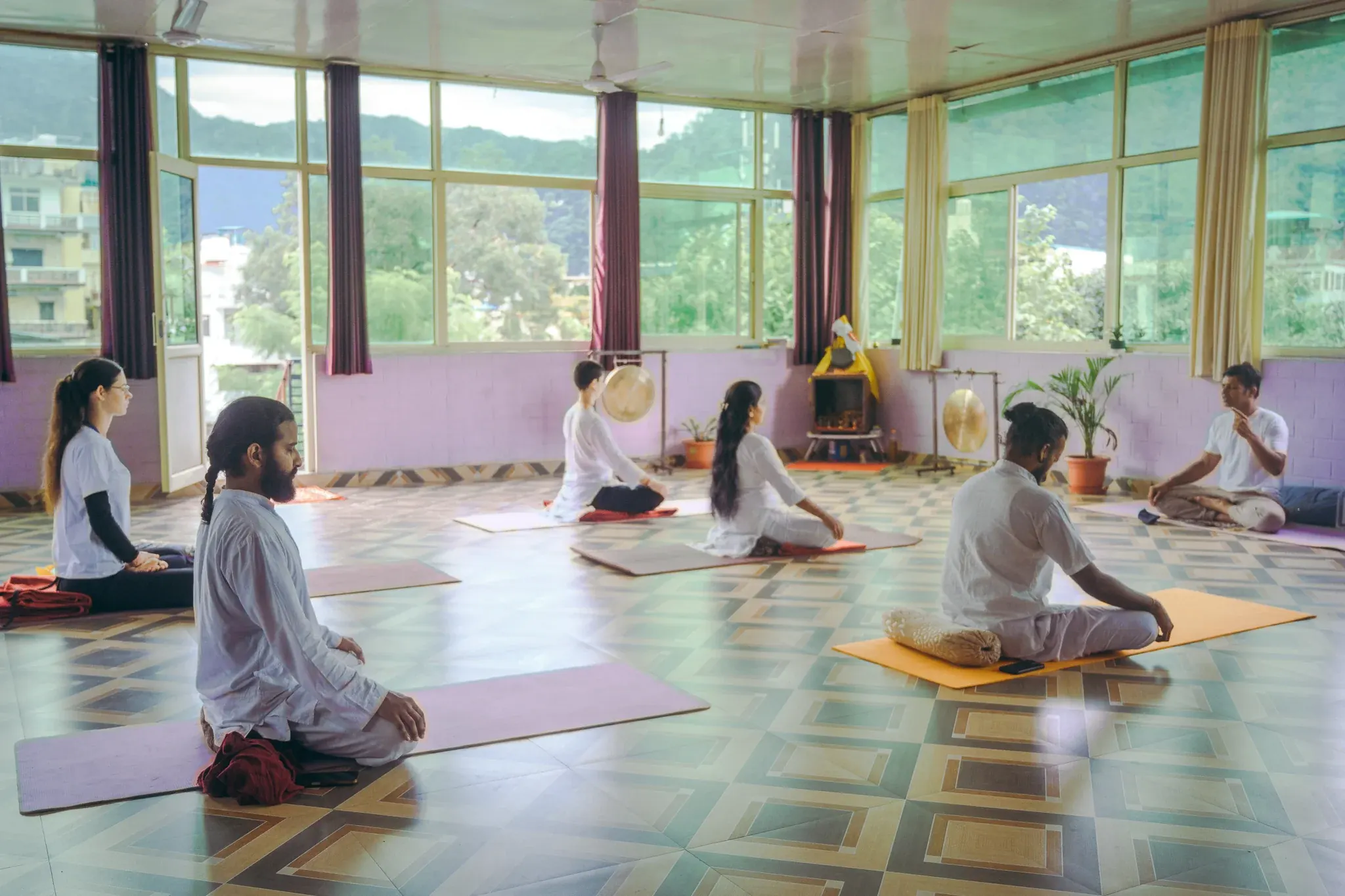 Teacher gesturing as he instructs students seated cross-legged in a bright Rishikesh shala facing the hills