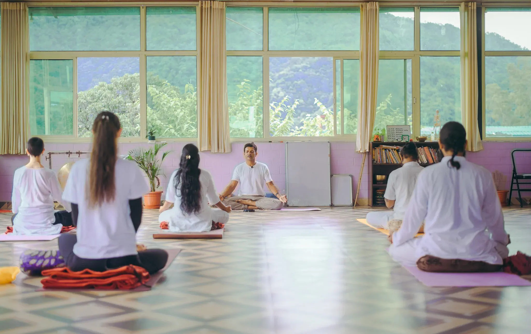 Teacher leading five seated students in a window-lined yoga shala facing green forested hills in Rishikesh